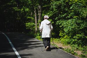 An elderly woman is leisurely walking along a road, aided by walking sticks, in a natural setting with plants, grass, and trees around her. She appears to be enjoying her stroll in nature photo
