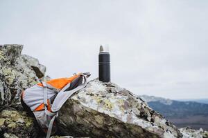 A thermos stands on a stone against the background of the sky in the mountains, a backpack lies on a rock, camping equipment, a halt in a mountainous area, photo