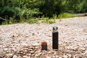 In a peaceful natural setting, there is a tranquil scene with a thermos and coffee cup placed on the ground among trees and grass, creating a sense of peace and serenity in the environment photo