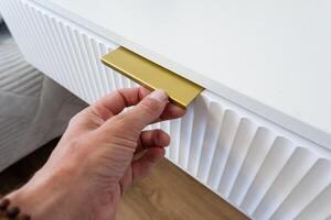 A person holds a shiny gold handle on a stylish white cabinet, matching the decor. The intricate design contrasts elegantly with the cabinets color, adding sophistication photo