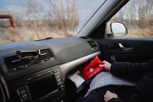 A person is seated comfortably inside a car, clutching a vibrant and easily identifiable bright red first aid kit, prepared to assist anyone in need during an emergency situation photo