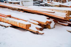 A substantial pile of wood, which is currently covered in a layer of snow, sits on a construction site, awaiting its future use in various building projects and structures photo