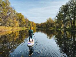 A man is riding a paddle board on a serene river, skillfully navigating the water while taking in the stunning natural scenery surrounding him, from lush trees to the open sky above photo