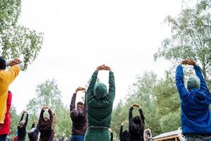 A diverse group of individuals stands in a circle, stretching their arms, relaxed and enjoying a leisurely outdoor event on grassy soil, surrounded by the beauty of nature under the sky photo
