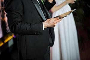 Man in tuxedo holds tablet while standing by bride and groom at formal event photo