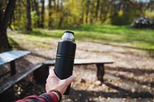 In a picturesque park teeming with vibrant greenery, a person stands proudly holding a thermos bottle in one hand, surrounded by lush trees and soft, inviting grass underfoot photo
