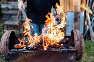 A man is cooking or working near a fire with a shovel amid flames and heat. The fire looks like it could be used for cooking or a similar activity, creating a warm atmosphere around him photo