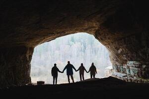 Silhouetted friends joyfully holding hands while standing at the captivating entrance of a cave, surrounded by beautiful nature scenery photo