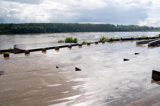 The scene depicts a river that has overflowed its banks, with water flooding the surrounding area. In the foreground, there is a dock, while trees are visible in the distant background photo