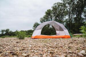 An empty camping tent is set up on a pebble beach with trees in the background on a cloudy day, creating a serene and picturesque landscape for outdoor enthusiasts to enjoy photo