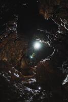 A solitary figure is standing inside a dimly lit cave, wearing a bright light on their head to illuminate the surrounding darkness, surrounded by various natural elements photo