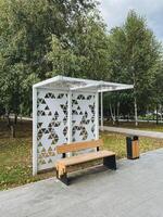 In a vibrant and lively public park, there is a bus stop that features a long comfortable bench along with a conveniently placed trash can for visitors waiting for their transportation photo