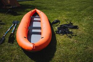 An orange inflatable kayak with paddles and gear is resting on green grass in a sunny park, suggesting outdoor recreation and water sports like kayaking and boating photo