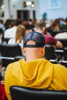 Man in yellow hoodie and blue hat sits in front of crowd at a competition event photo