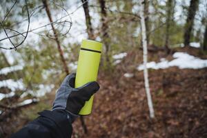 A green thermos on the background of a spring forest, a man holds a thermo bottle with a gloved hand. photo