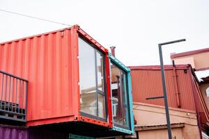 A vibrant red shipping container, which features a balcony, is placed alongside a building, creating an interesting contrast in colors and materials, while enhancing the urban landscape photo