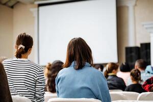 Audience watching a presentation in a seminar room with a white screen background. People at a formal educational event, listening to a speaker in a business setting photo