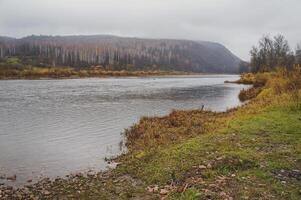 Water flows by a mountain, trees line the shore under a cloudy sky photo