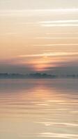 Serene Sunrise Over Calm Water With Soft Reflection And Misty Horizon Silhouetting Trees photo