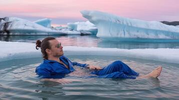 Man Relaxing in Arctic Ice Pool Wearing Blue Shirt with Icebergs and Pink Sunset Sky Reflection photo