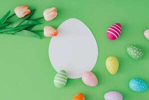 Spring composition with colorful Easter eggs and tulip flowers on pastel green background with paper card note copy space. Minimal Easter concept. Creative festive holiday flat lay. Easter aesthetic. photo