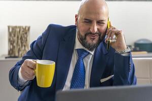 portrait of smiling businessman talking on mobile phone in front of laptop during coffee break in an office photo