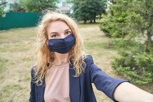 Caucasian young female in mask taking selfie in outdoor park setting with trees in background. photo