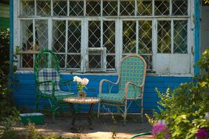 Wicker chairs against background of old country house near round table on which there is glass vase with white flowers inside. photo