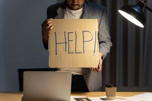 Young businessman working on computer laptop asking for help holding cardboard sign looking desperate and depressed in stress overwhelmed. photo