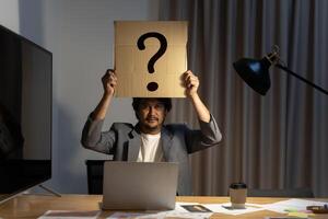 Young man with a question sign on a card covering face. Employee's dilemma with question mark on blank paper. Confused, Thinking, Brainstorming or Decision. photo