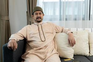 Portrait of handsome young Arab man smiling and looking at camera, sitting on sofa at home. photo