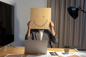 A young businessman is holding a cardboard with a smiley face on it in front of his head, gesturing with a cardboard box. photo