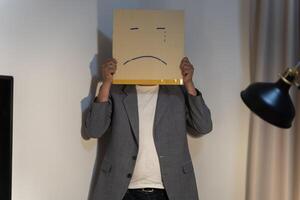 Unhappy man employee with box instead of his head. Portrait of businessman covering his face with paper mask with sad face drawn on it. photo