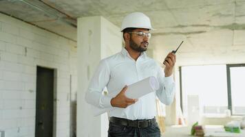 Indian civil engineer stands at a construction site holding blueprints and communicating via walkie-talkie video
