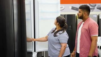 Indian couple open door of the refrigerator in the appliances store and compare with other models to buy the new house video