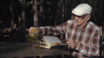 An elderly man in a flat cap pours tea into a cup while seated at a wooden table, immersed in a book amidst a serene forest backdrop on a sunny afternoon. video