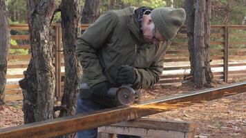 A person wearing warm clothing operates a grinder on a metal beam, creating sparks in a tranquil forested area during daylight. Trees surround the workspace. video