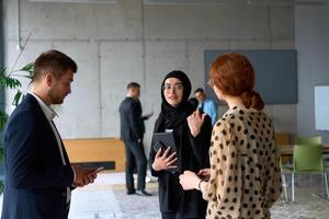 A diverse group of business professionals engages in a conversation during a work break in the office, with their colleagues visible in the background, fostering collaboration and connection photo