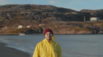 Attractive man, wearing yellow raincoat and red hat, steps forward and gazes into camera. He fisherman or an employee of seaport. Behind him, there are rocks, tundra, and ocean. video