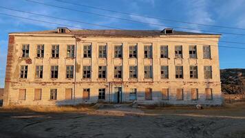 An old, abandoned school, with boarded-up and broken windows on the ground floor, stands in the middle of harsh, northern landscape on bright, sunny day. video
