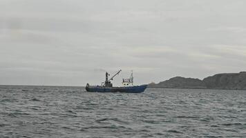 Fishing trawler sets sail to Arctic Ocean in search of fish, with backdrop of colorless, steep cliffs beyond Arctic Circle. Slow motion. video