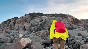 Traveler, clad in vibrant yellow raincoat, and equipped with backpack, begins ascent of hill, which is covered in diverse rocks and boulders. video