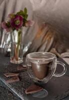 winter still life with glass transparent cup of hot chocolate on table surrounded by chocolate pieces and black chia seeds on background hellebore flowers,desert concept in color of 2025,mocha mousse photo
