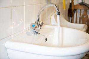 Flowing water from a shiny chrome faucet into a white sink, tiled wall background, clean and modern design concept, photo