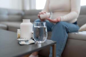 Unrecognizable anonymous pregnant woman holding jar with pills taking vitamins and minerals treating sickness during pregnancy sitting on sofa at home photo