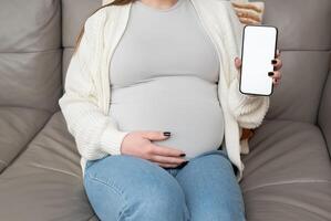 Close-up of pregnant woman holding and showing mobile phone with empty display at home. Mockup image with copy space for app or interface customization photo
