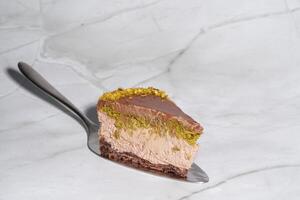 Slice of chocolate Dubai cheesecake with pistachio paste and kataifi covered in chocolate ganache on cake spatula. White background with copy space. Indulgence, gourmet desserts, culinary presentation photo