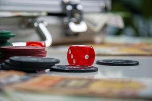 A red dice sits on a table with black and red chips, a stack of playing cards, and a blurred background photo