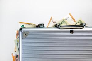 A silver metal briefcase sits closed with US dollar and euro bills partially visible inside photo