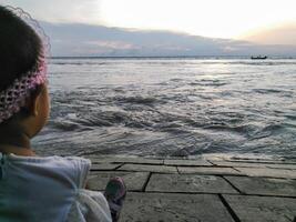 A serene sunset over a body of water with a person sitting on a paved area near the water. A boat with two people is visible on the horizon, and the partly cloudy sky adds to the tranquil atmosphere. photo